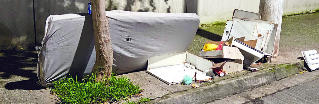Colchão descartado na rua, encostado em um muro atrás de uma árvore, à noite